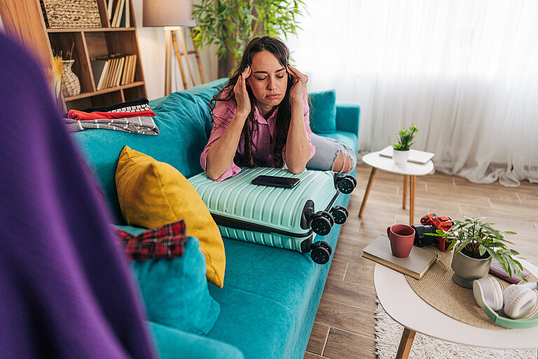 Frau lehnt gestresst auf ihrem gepackten Koffer und reibt sich die Schläfen.