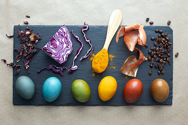 Eier in verschiedenen Farben. Die natürlichen Produkten, mit welchen das entsprechende Ei gefärbt wurde, liegen darüber.