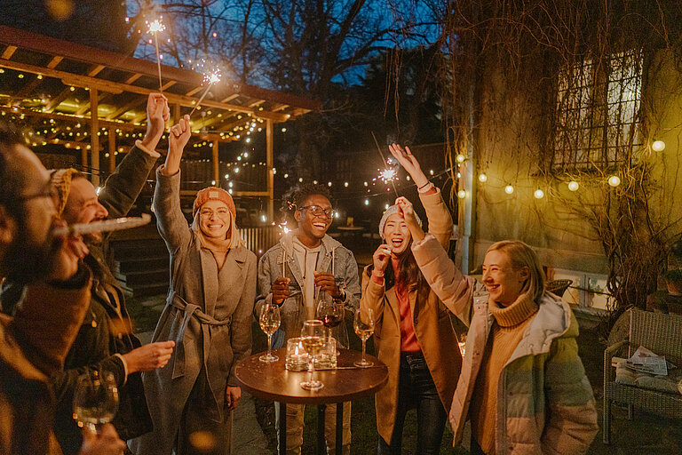 Freundesgruppe feiert Silvester draußen, stehen um einen Stehtisch mit Weingläsern darauf und halten Wunderkerzen nach oben.