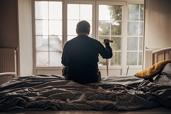 Älterer Mann mit Gehstock sitzt allein auf seinem Bett und schaut nach draußen. 