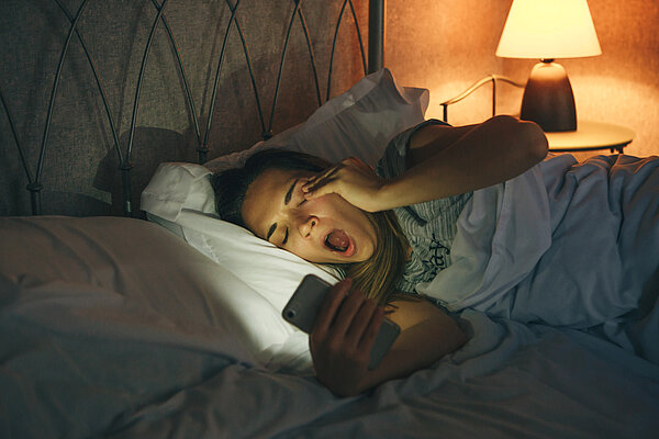 Frau liegt abends im Bett und schaut aufs Smartphone. Dabei gähnt sie. 