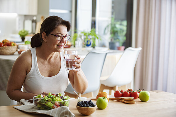 Frau trinkt Wasser und hat wasserreiche Lebensmittel (Obst und Gemüse) vor sich auf dem Tisch.