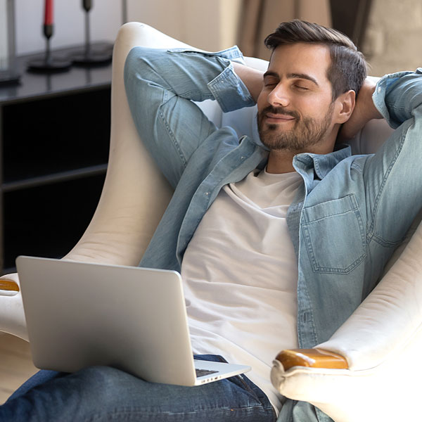 Ein Mann sitzt zurückgelehnt in einem Sessel mit Laptop auf dem Schoß. Hat die Augen geschlossen und lächelt.