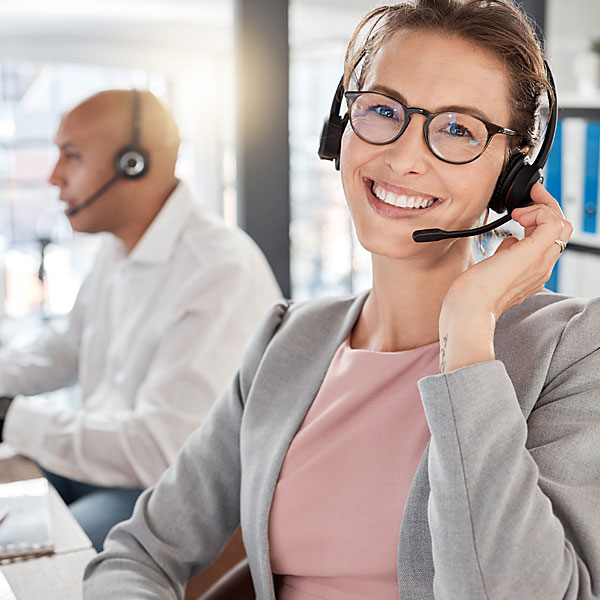 Frau mit Headset lächelt, Mann mit Headset im Hintergrund. 