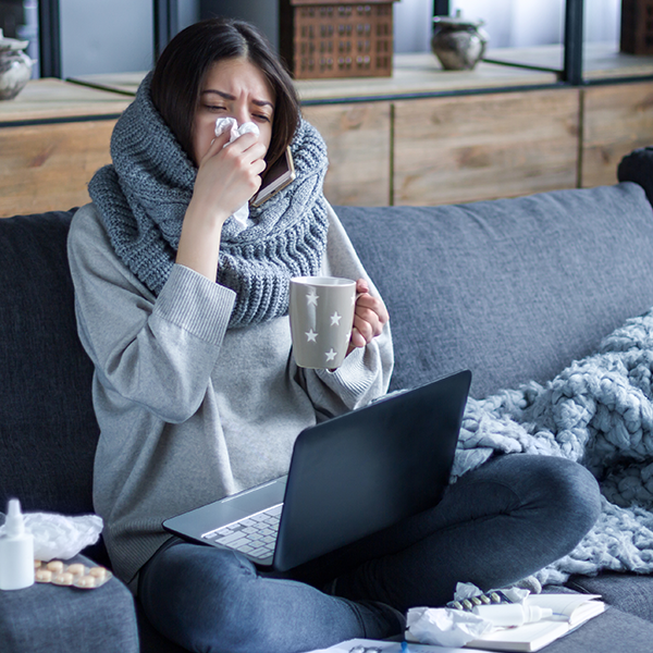 Kranke Frau mit dickem Schal und Tasse in der Hand sitzt auf dem Sofa mit Laptop auf dem Schoß und putzt sich die Nase.
