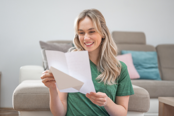 Eine Frau sitzt zu Hause vor dem Sofa und freut sich über einen Brief den sie in der Hand hält