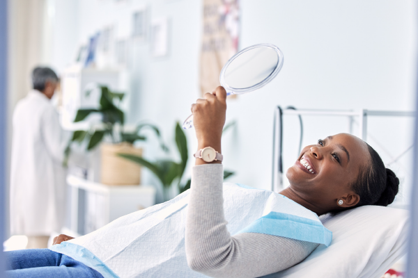 Eine Frau liegt beim Zahnarzt auf dem Behandlungsstuhl und schaut sich glücklich das Ergebnis im Spiegel an. 